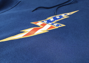 Close up chest region view of the navy blue Freedom Bolt hoodie, showcasing a gold outlined 7-bolt with white stars on blue on top and red and white stripes on the bottom. 