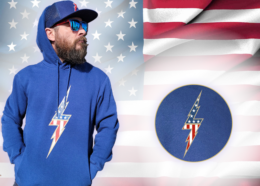 Full view of the navy blue Freedom Bolt hoodie, featured on a male model also sporting the 7-Strong trucker hat and showcasing a gold outlined 7-bolt with white stars on blue on top and red and white stripes on the bottom. The hoodie is featured on an American flag background. There is a detail circle in the bottom right circle featuring the 7-Bolt on the hoodie