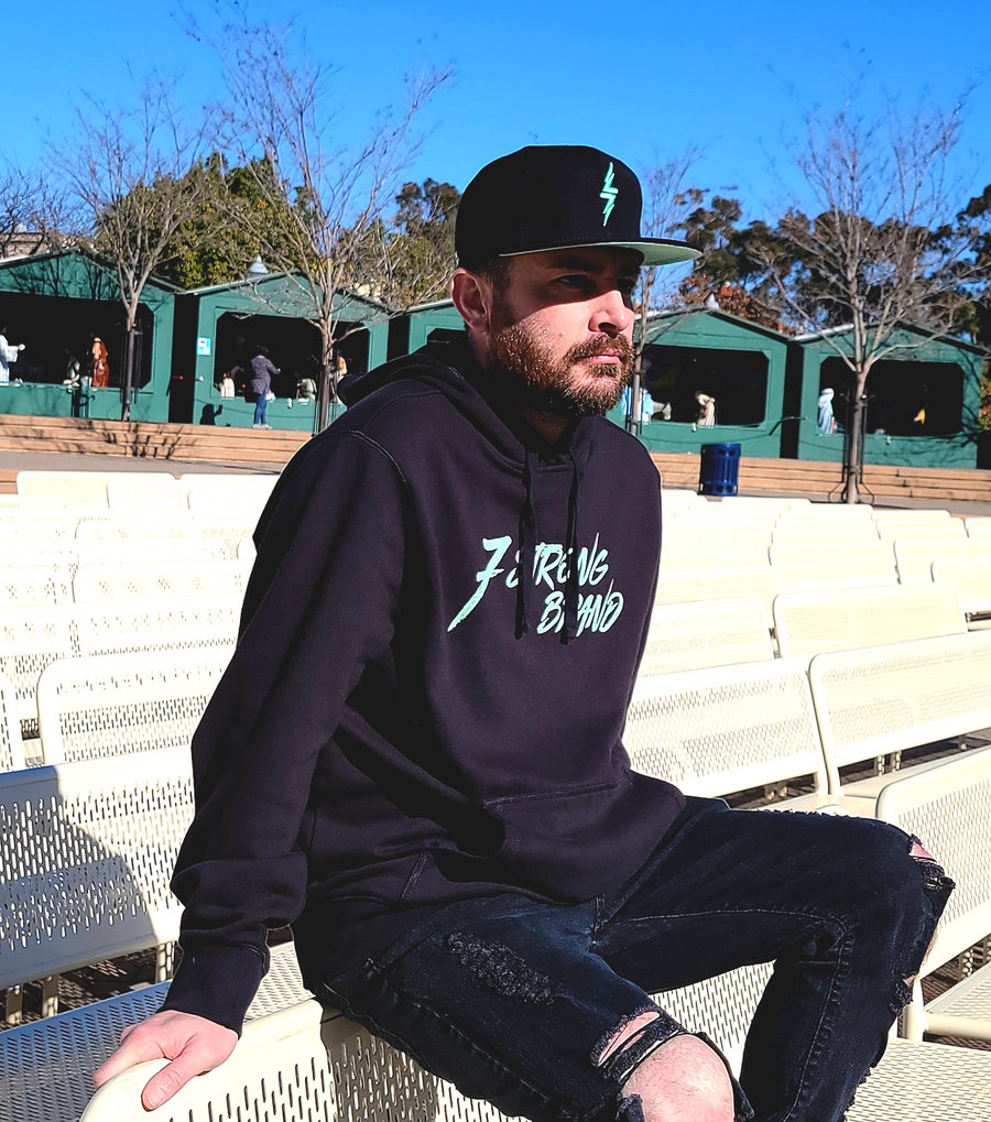 Model sporting the Black snapback hat on a futuristic background, hat features the mint version of the 7-Strong bolt logo in the park, along with the 7-Strong script branded hooded sweatshirt. 