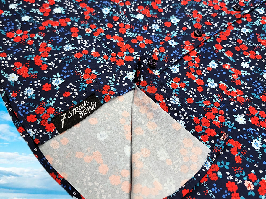 3/4 view of sweep tag portion on the 7-Strong "Aye Poppy" button down, featuring an array of red poppys with white sprigs on a deep navy blue shirt. The shirt is displayed against a partly cloudy sky. 