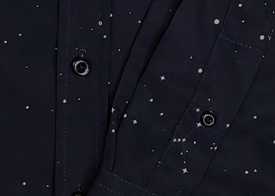 Midsection and draping sleeve view of the long-sleeve Stargazer button down shirt. A deep navy blue shirt with constellation star patterns throughout. The shirt is displayed against a night sky full of stars. 
