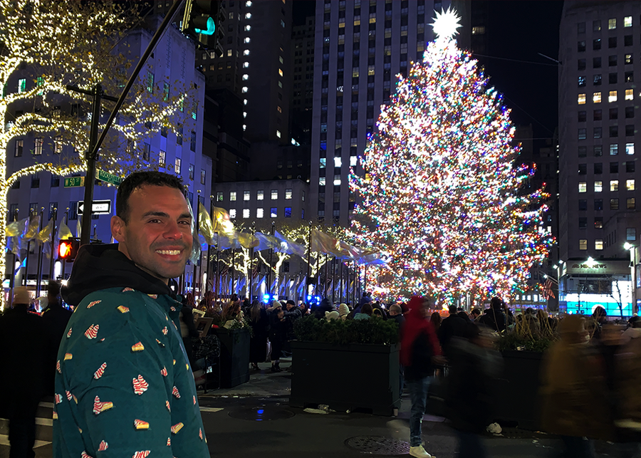 Male model in front of Christmas Tree in a city setting, smiling back to camera sporting the long-sleeve "Oh, Christmas Treat" shirt - a green, Christmas sweater-like background with columns of various white Christmas tree shaped cakes with red garland, some whole, some bitten. 