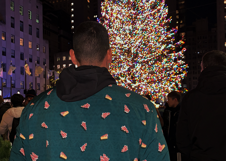 Adult male model wearing long-sleeve "Oh, Christmas Treat" shirt with a green, christmas sweater-like background with columns of various white Christmas tree shaped cakes with red garland, some whole, some bitten. Model is standing in front of a Christmas Tree. 