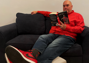 Male model sitting on a couch in red and black shoes, reading a book while wearing the 7-Strong Red 7-Bolt long sleeve shirt, featuring black 7-bolts with a white drop shadow interspersed on a red background. 