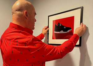 Medium shot of a male model framing a red and black sneaker photo on wall while wearing the 7-Strong Red 7-Bolt short sleeve shirt, featuring black 7-bolts with a white drop shadow interspersed on a red background.