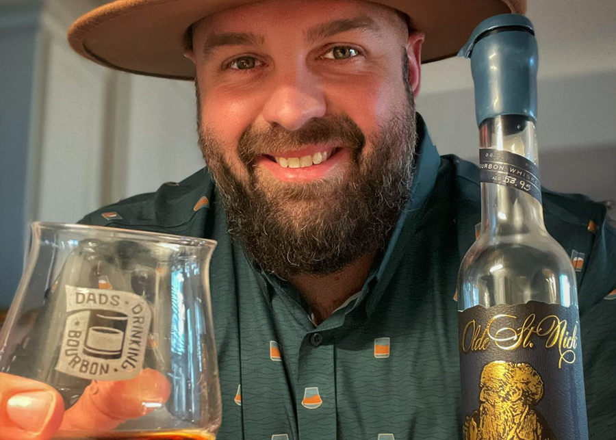 Male model, drinking a bourbon, smiling at camera and wearing the 7-Strong "Straight Sippin'" long Sleeve button down. The shirt is a deep green with black ripples as its background, while the foreground has various drinking glasses with various levels of whiskey, some with ice, others without.