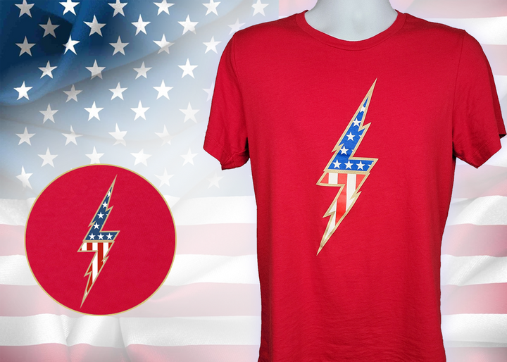 Full view of the red Freedom Bolt unisex tee, featuring a large center placed 7-bolt, outlined in gold, with white stars on blue on top and red and white stripes on the bottom. The shirt is featured on a mannequin against a U.S. flag background. In the bottom left corner, there is a detail circle featuring an up close view of the Freedom Bolt.