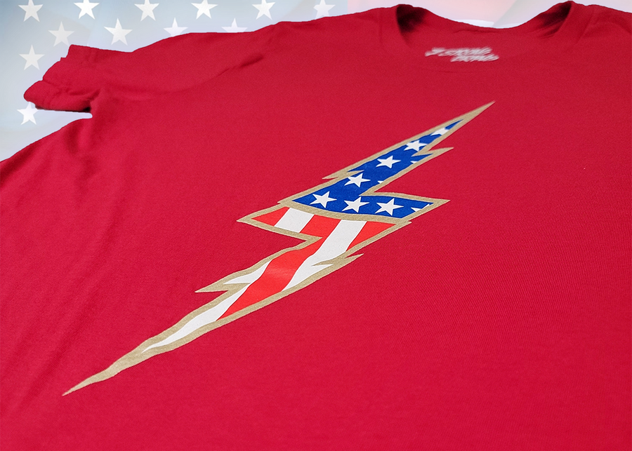 Angle middle view of the red Freedom Bolt unisex tee, featuring a large center placed 7-bolt, outlined in gold, with white stars on blue on top and red and white stripes on the bottom. The shirt is featured on a U.S. flag background.