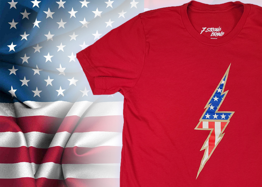 3/4 view of the red Freedom Bolt unisex tee, featuring a large center placed 7-bolt, outlined in gold, with white stars on blue on top and red and white stripes on the bottom. The shirt is featured on a  U.S. flag background. 