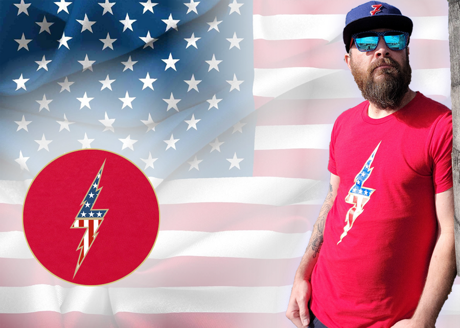 Full view of the red Freedom Bolt unisex tee, featuring a large center placed 7-bolt, outlined in gold, with white stars on blue on top and red and white stripes on the bottom. The shirt is featured on a medium male model against a U.S. flag background. In the bottom left corner, there is a detail circle featuring an up close view of the Freedom Bolt.