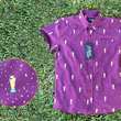 Load image into Gallery viewer, Full view of the Cup of Life youth button up, a off-maroon colored shirt decorated throughout with falling gold confetti and depictions of the World Cup statue. The shirt is laid out on a grass pitch. In the bottom left corner is a detail circle, highlighting the shirt&#39;s design.
