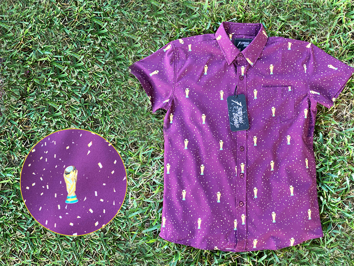 Full view of the Cup of Life youth button up, a off-maroon colored shirt decorated throughout with falling gold confetti and depictions of the World Cup statue. The shirt is laid out on a grass pitch. In the bottom left corner is a detail circle, highlighting the shirt's design.