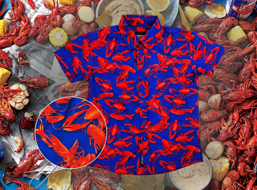 Full view of the youth 7-Strong Dat Boil shirt, with design detail circle in bottom left corner, in deep navy blue with red crawfish patterned throughout overlapping one another. The shirt itself sits on a background image of items from a Crawfish Boil such as crawfish, seasoning, corn, and potatoes. 