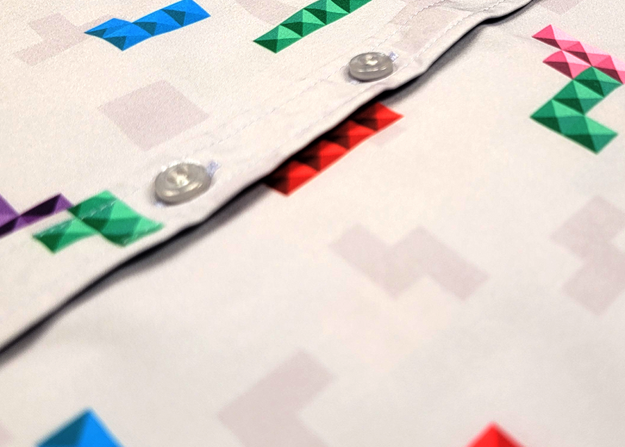 Close up of the middle button portion of the Block Party shirt detailing the various colored blocks of different arrangements inspired by a classic video game, cascading down the shirt against a grayish background featuring shadows of the same shapes. The shirt is presented on a background of similar design.