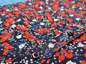 Middle button region view of the youth 7-Strong "Aye Poppy" button down, featuring an array of red poppys with white sprigs on a deep navy blue shirt. The shirt is displayed against a partly cloudy sky. 
