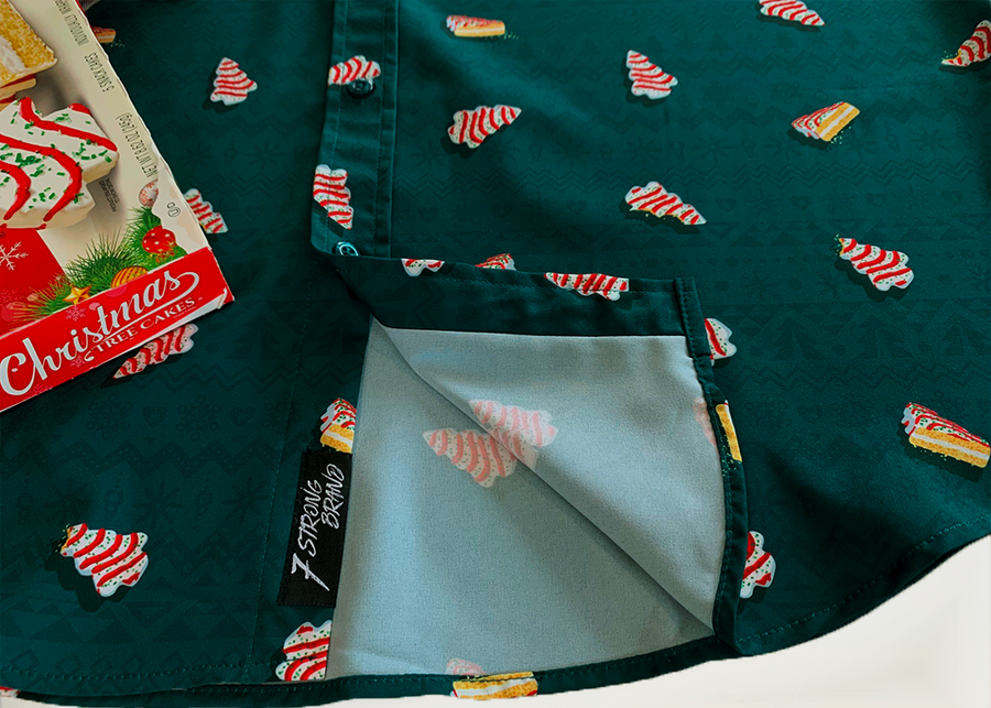 Bottom, sweep tag portion view of the 7-Strong "Oh, Christmas Treat" shirt - a green, christmas sweater-like background with columns of various white Christmas tree shaped cakes with red garland, some whole, some bitten. The shirt is displayed against a red, snowing background with a similar parted cake. 