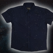 Load image into Gallery viewer, Full view of the short-sleeve youth Stargazer button down shirt. A deep navy blue shirt with constellation star patterns throughout. The shirt is displayed against a night sky full of stars.
