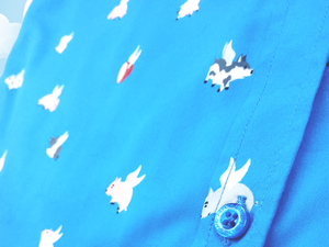 Close Up shot of the midsection area of the 7-Strong "When Pigs Fly" shirt, showcasing various pigs and meats flying against a blue, muted cloud background. Shirt is displayed against a background of cartoon clouds.