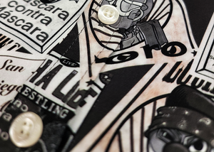 Midsection button view of the youth 7-Strong "Cinco de Mayhem" button down shirt. Shirt features black and white posters featuring various created luchadors advertised for matches, all overlapping each other over a black shirt. Shirt is displayed on a black gradient background with some of the characters ghosted in the back.