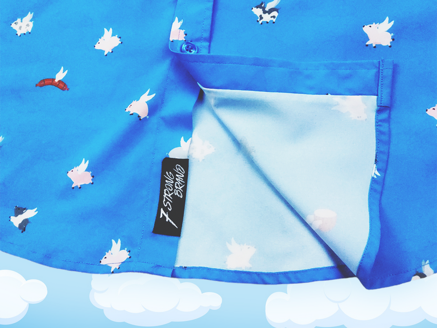 Close up shot of sweep tag region of the 7-Strong "When Pigs Fly" shirt, showcasing various pigs and meats flying against a blue, muted cloud background. Shirt is displayed against a background of cartoon clouds.