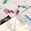 Load image into Gallery viewer, Close up of the bottom, sweep tag, portion of the Block Party shirt detailing the various colored blocks of different arrangements inspired by a classic video game, cascading down the shirt against a grayish background featuring shadows of the same shapes. The shirt is presented on a background of similar design.

