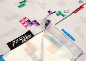Close up of the bottom, sweep tag, portion of the Block Party shirt detailing the various colored blocks of different arrangements inspired by a classic video game, cascading down the shirt against a grayish background featuring shadows of the same shapes. The shirt is presented on a background of similar design.