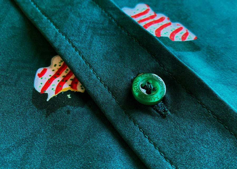 Close up, mid-button view of the 7-Strong "Oh, Christmas Treat" shirt - a green, christmas sweater-like background with columns of various white Christmas tree shaped cakes with red garland, some whole, some bitten. The shirt is displayed against a red, snowing background with a similar parted cake. 