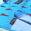 Load image into Gallery viewer, View of the bottom sweep tag portion of youth 7-Strong &quot;Pennant to Win It&quot; shirt, which is sky blue with texture, featuring various triangular flags featuring baseball&#39;s prominent cities patterned throughout. The shirt is featured against abackground is sunset at a major league stadium. 
