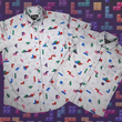 Load image into Gallery viewer, Full view of the 7-Strong Block Party adult and youth button downs overlapping each other, featuring various colored blocks of different arrangements inspired by a classic video game, cascading down the shirt against a grayish background featuring shadows of the same shapes. The shirt is presented on a background of similar design.

