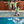 Load image into Gallery viewer, Youth model wearing the 7-Strong &quot;Late Bloomer&quot; youth button down - a yellow background shirt with white and blue flowers patterned all over it. Model is being shown multiple times in edited phases of jumping into a pool with the shirt on and googles... from beginning to jumping in to splashing into the pool. 

