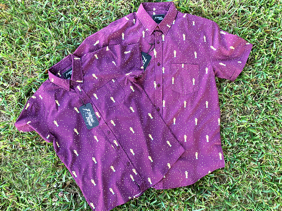 Full view of the Cup of Life adult and youth button ups, overlapping one another, a off-maroon colored shirt decorated throughout with falling gold confetti and depictions of the World Cup statue. The shirt is laid out on a grass pitch. 
