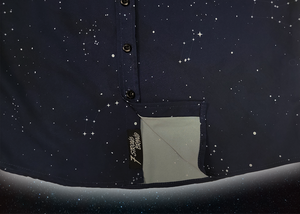 Bottom sweep tag portion view of the short-sleeve youth Stargazer button down shirt. A deep navy blue shirt with constellation star patterns throughout. The shirt is displayed against a night sky full of stars.