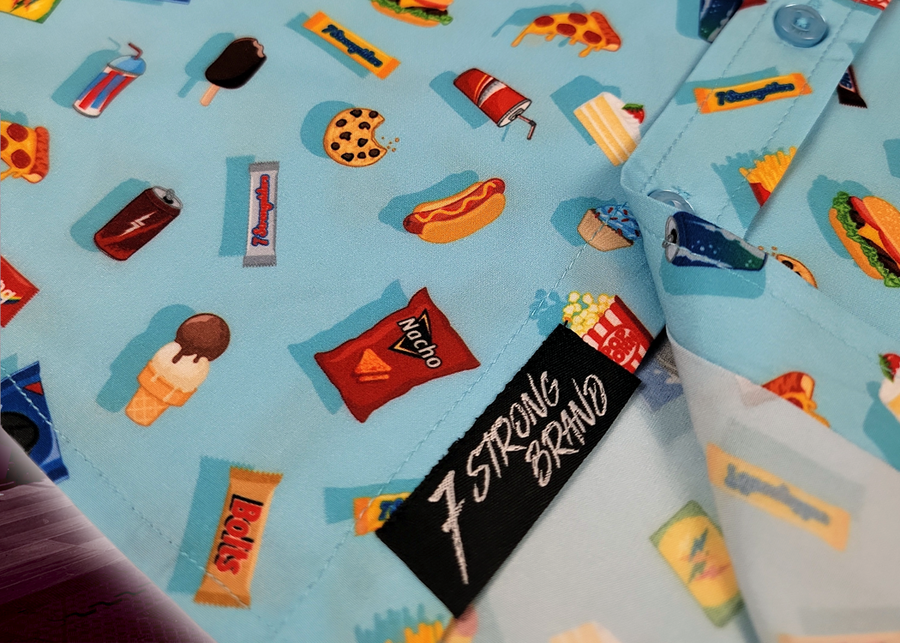 Bottom sweep tag view of the 7-Strong "Cheat Day" youth button down featuring various snacks, treats, and fast food features on a light blue shirt. The shirt is displayed against a background of a gym weight rack
