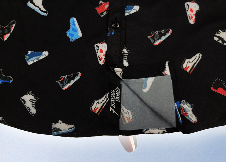 Bottom sweep tag view of the youth short sleeve version of "Got 'Em!" The button down features various athletic fashion sneakers in an 80bit format arranged all over a black shirt. The shirt is featured against a background of a basketball player jumping in air, noticeable sneakers being worn. 
