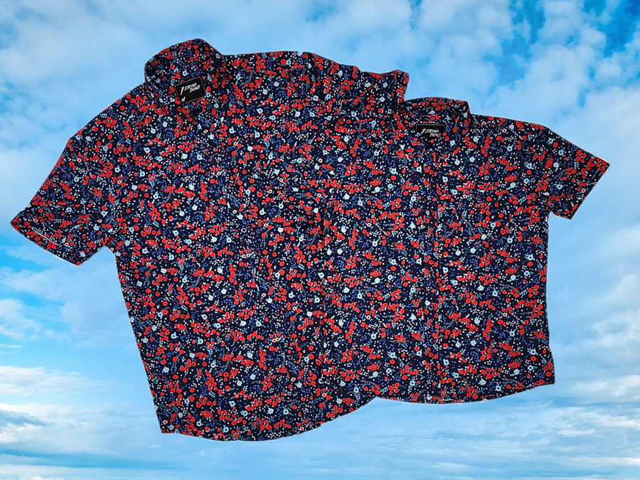 Full, overlapping view of the adult and youth 7-Strong "Aye Poppy" button down, featuring an array of red poppys with white sprigs on a deep navy blue shirt. The shirt is displayed against a partly cloudy sky. 