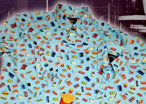 view of the 7-Strong "Cheat Day"  adult and youth button downs overlapping each other featuring various snacks, treats, and fast food features on a light blue shirt. The shirt is displayed against a background of a gym weight rack