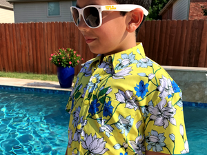 Youth model wearing the 7-Strong "Late Bloomer" youth button down - a yellow background shirt with white and blue flowers patterned all over it. Model is standing by the pool in sunglasses with the shirt being shown at a medium close up on the body. 