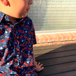 Load image into Gallery viewer, Male youth model sitting on a deck looking off in the distance wearing the youth &quot;Aye Poppy&quot; button down shirt displaying red poppies with white sprigs on a deep navy blue background. 
