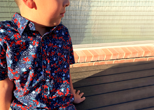 Male youth model sitting on a deck looking off in the distance wearing the youth "Aye Poppy" button down shirt displaying red poppies with white sprigs on a deep navy blue background. 