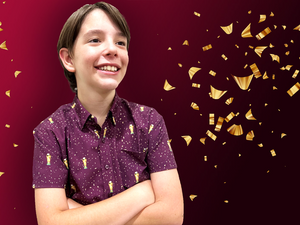 Youth male model, arms crossed and smiling, sporting the Cup of Life adult and youth button up, a off-maroon colored shirt decorated throughout with falling gold confetti and depictions of the World Cup statue. The models are standing against a confetti background that matches the shirt design.
