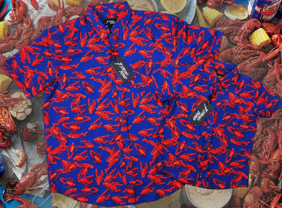 Full view of the both the youth and adult 7-Strong Dat Boil shirts side-by-side in deep navy blue with red crawfish patterned throughout overlapping one another. The shirt itself sits on a background image of items from a Crawfish Boil such as crawfish, seasoning, corn, and potatoes. 