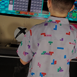 Load image into Gallery viewer, Medium shot of youth male in the 7-Strong Block Party adult button down, featuring various colored blocks of different arrangements inspired by a classic video game, cascading down the shirt against a grayish background featuring shadows of the same shapes. Model is playing a video game, back turned to camera. 
