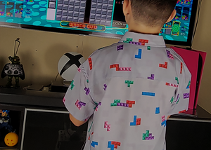 Medium shot of youth male in the 7-Strong Block Party adult button down, featuring various colored blocks of different arrangements inspired by a classic video game, cascading down the shirt against a grayish background featuring shadows of the same shapes. Model is playing a video game, back turned to camera. 