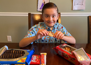 Child model wearing the 7-Strong "Cheat Day" youth button down featuring various snacks, treats, and fast food features on a light blue shirt indulging in some cookies and candy. 