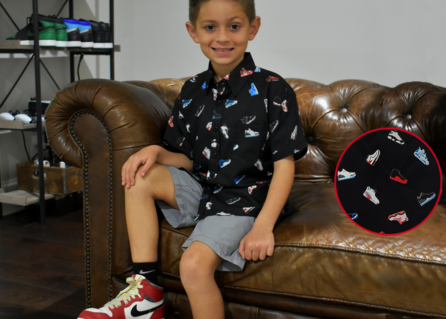 Male youth modeling the youth short sleeve version of "Got 'Em!" The button down features various athletic fashion sneakers in an 8-bit format arranged all over a black shirt.  Child is sitting on a coach looking at camera, while on the right is detail circle showcasing various shoes designed on the shirt. 