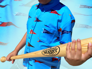 Midsection view of a youth male model, yielding baseball bat, sporting the 7-Strong "Pennant to Win It" shirt, which is sky blue with texture, featuring various triangular flags featuring baseball's prominent cities patterned throughout. The shirt is featured against a background is a backdrop of the Pennants featured on the shirt. 