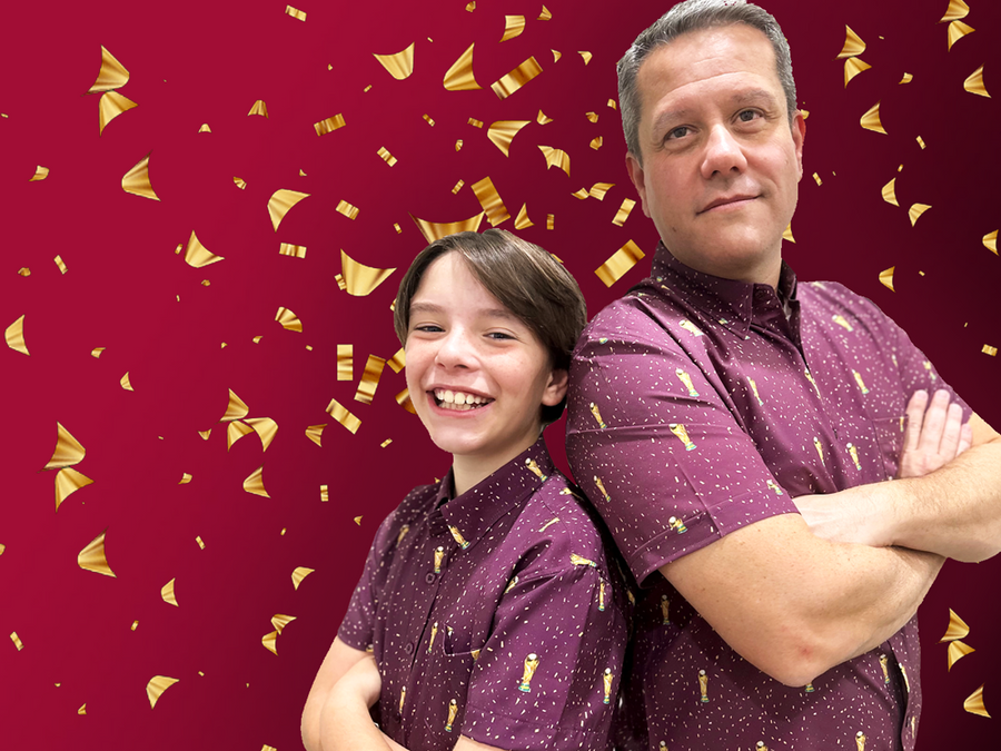 Male models, standing back-to-back, sporting the Cup of Life adult and youth button up, a off-maroon colored shirt decorated throughout with falling gold confetti and depictions of the World Cup statue. The models are standing against a confetti background that matches the shirt design.