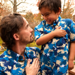Load image into Gallery viewer, Medium close up male adult model and youth model with their hands over their hearts, as if saluting the flag, looking at each other and sporting the 7-Strong &quot;Service Stars&quot; adult and youth button-ups, featuring blue and creme colored camouflage with white weathered stars throughout. 
