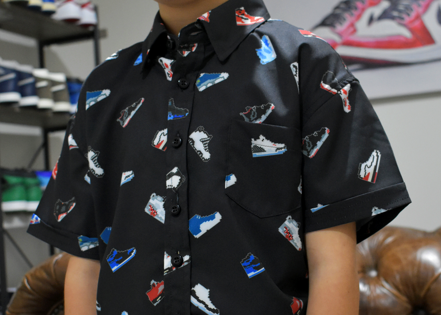 Midsection close up of male child model wearing the youth short sleeve version of "Got 'Em!" The button down features various athletic fashion sneakers in an 80bit format arranged all over a black shirt. The model is standing in a room with visible sneakers displayed behind him. 