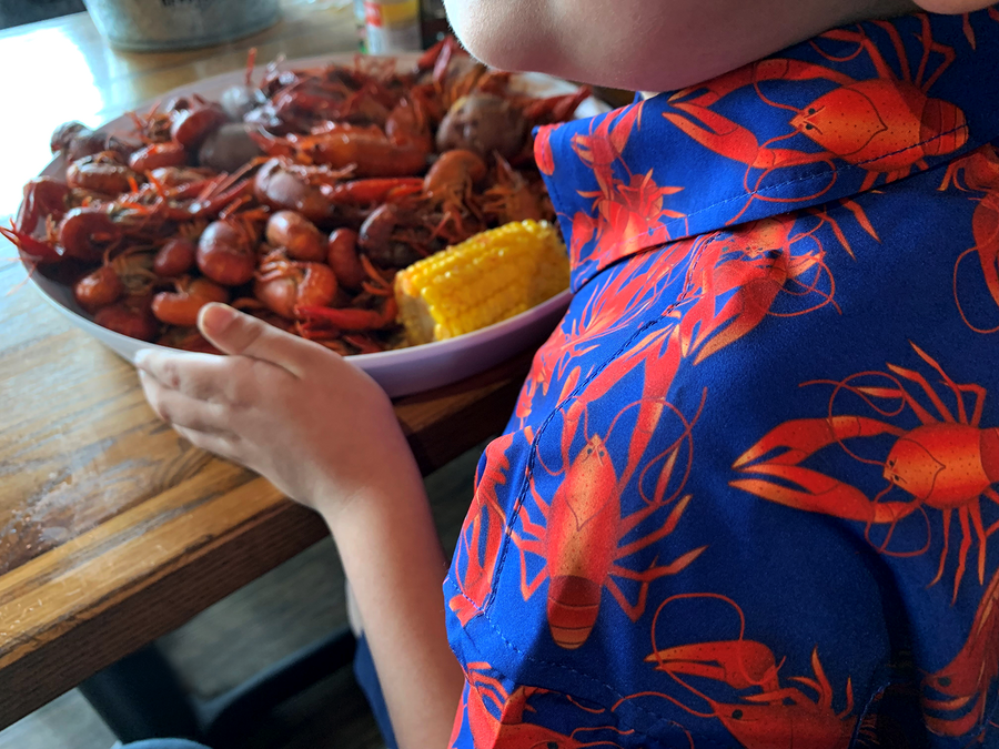 Close up, behind the shoulder view of model wearing the youth 7-Strong Dat Boil shirt in deep navy blue with red crawfish patterned throughout overlapping one another. The model is sitting in front of a bowl of crawfish with a corn cob. 